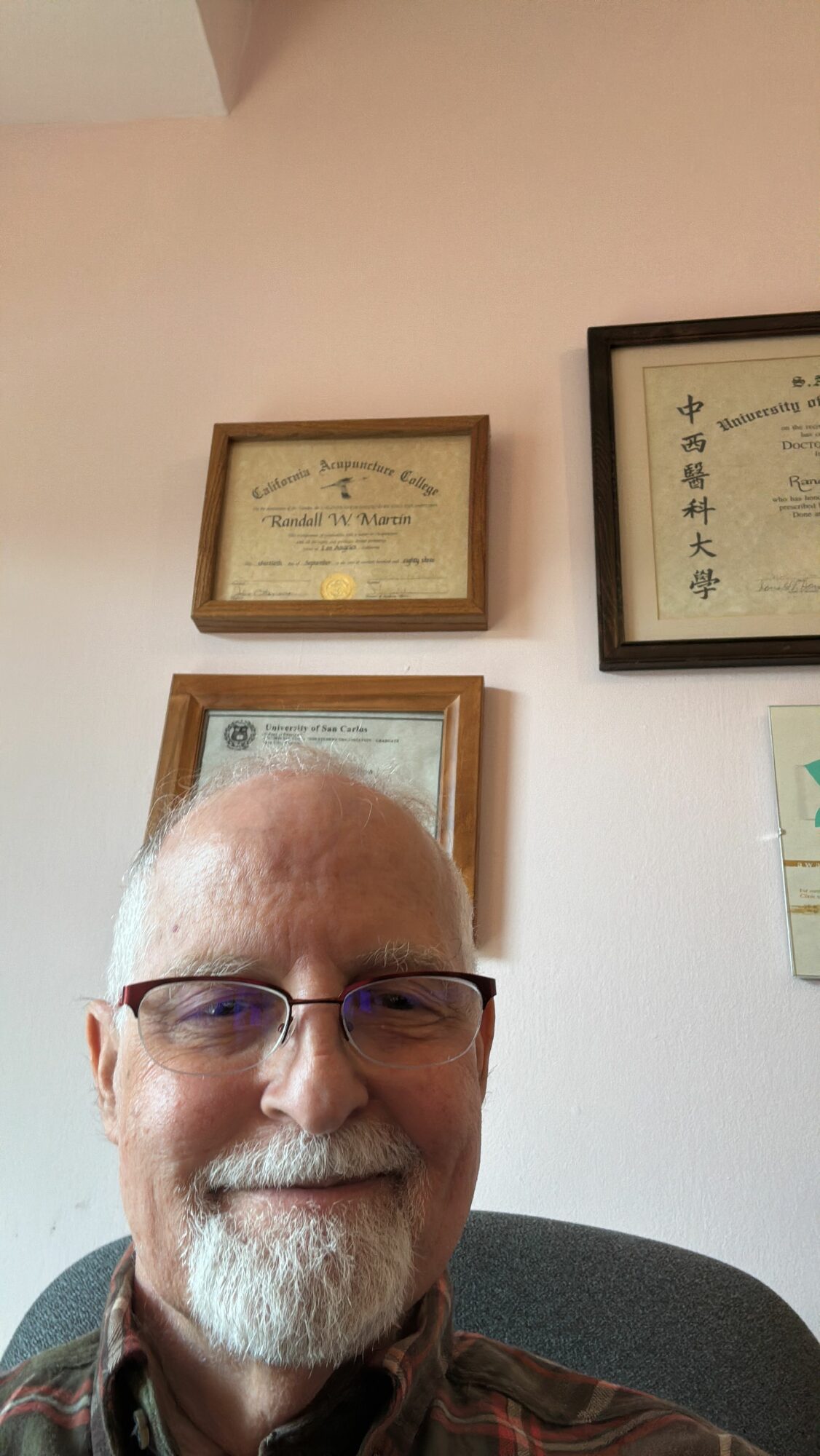 An elderly man with glasses and a beard, smiling, sitting at a table in a room with framed pictures on the wall.