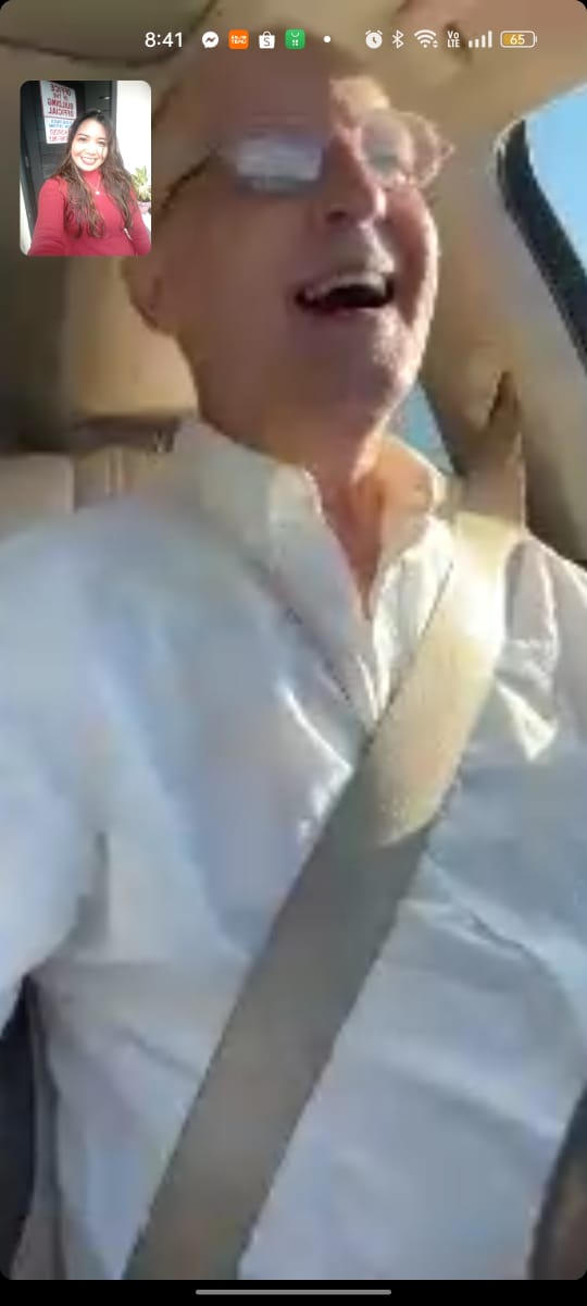 Older man with glasses and white shirt smiling while driving, with a woman in a red top in a small inset photo.