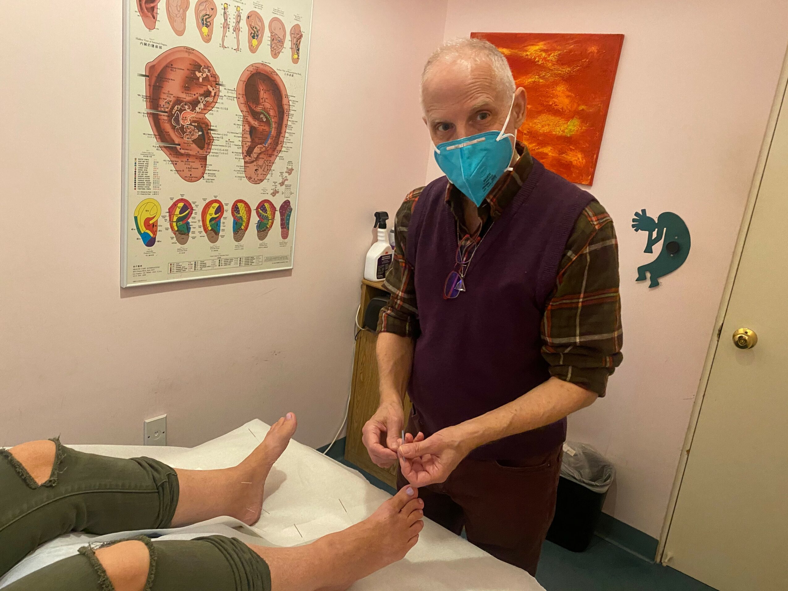 Person wearing a blue face mask providing foot care to a patient lying on a bed, with medical posters on the wall.