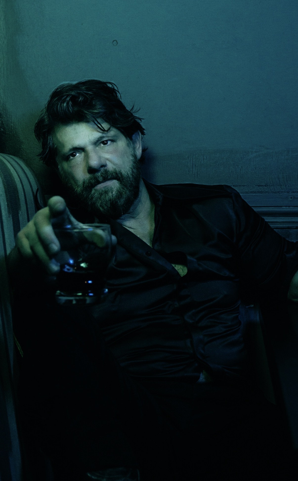 Man with beard and wavy hair sitting in a dimly lit room, holding a glass, looking at the camera.