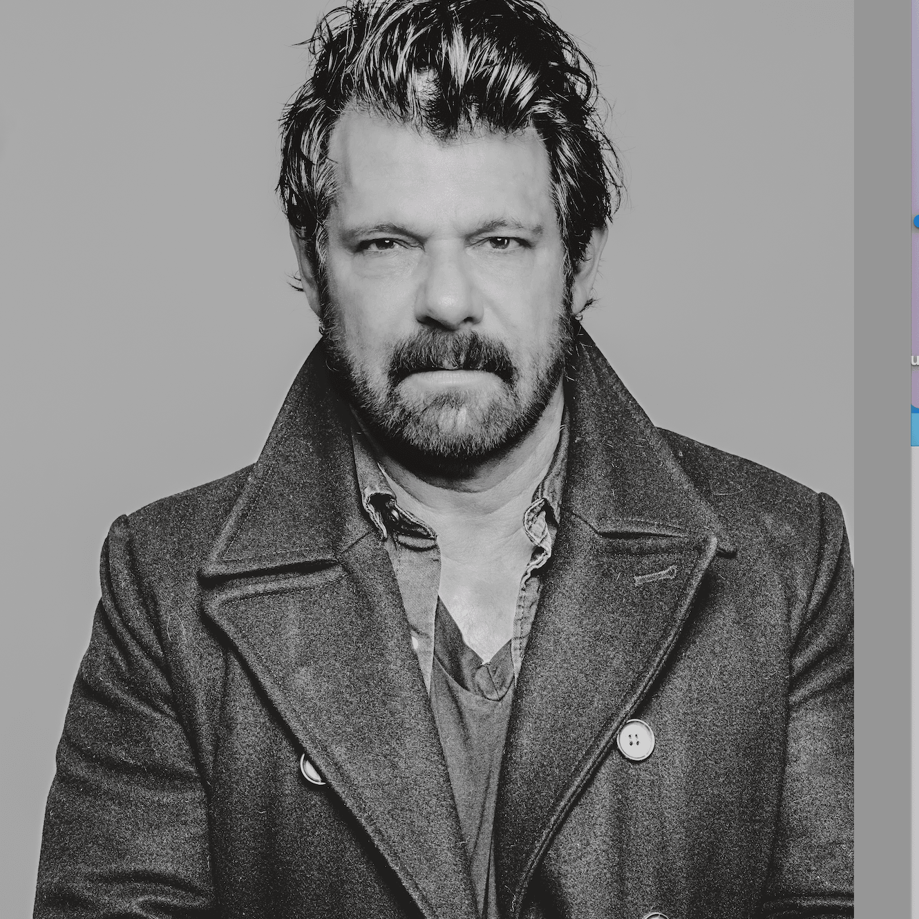 Man with beard and tousled hair wearing a coat and shirt, looking serious, against a plain background.
