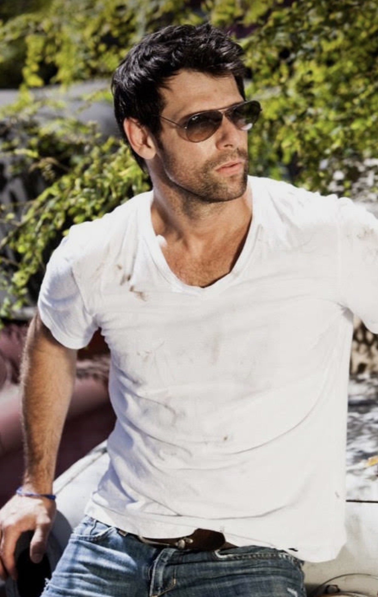 Man with dark hair and sunglasses wearing a white t-shirt outdoors, with greenery in the background.