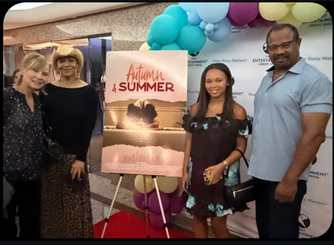 Four people standing outdoors near a colorful balloon display and a poster for Autumn & Summer, smiling at the camera.