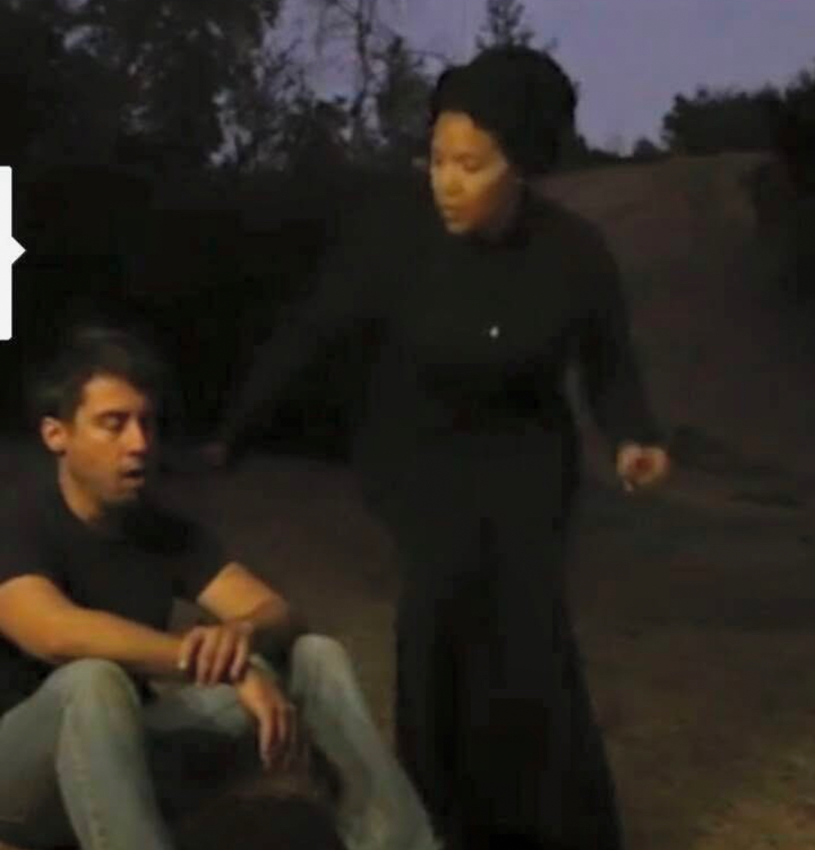 A woman standing next to a seated man outdoors during dusk, with trees in the background.
