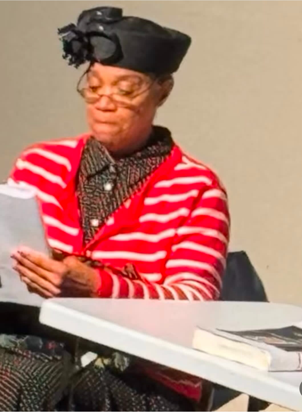 Woman wearing glasses, a black hat, and a red and white striped sweater, reading a paper at a table.