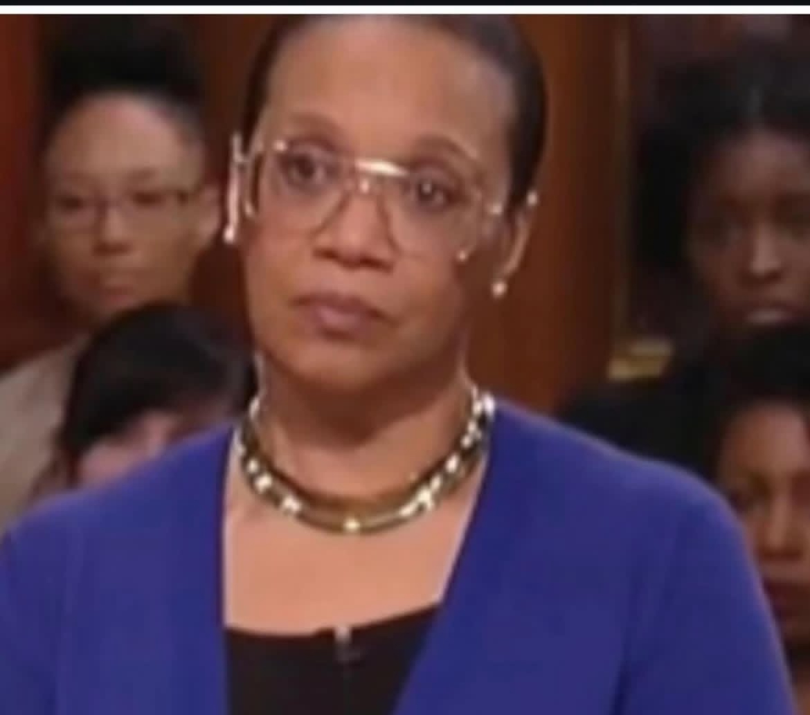 Woman with glasses and necklace, wearing a blue blazer, in a room with other people in the background.
