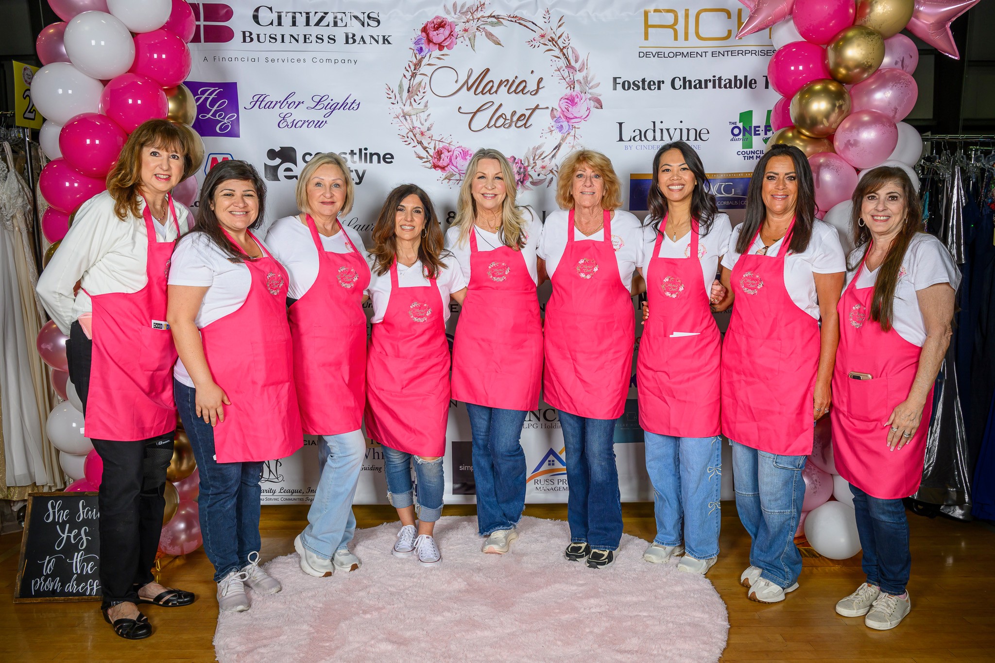 Group of nine women wearing pink aprons and white shirts, standing in front of a decorated backdrop with balloons and banners.