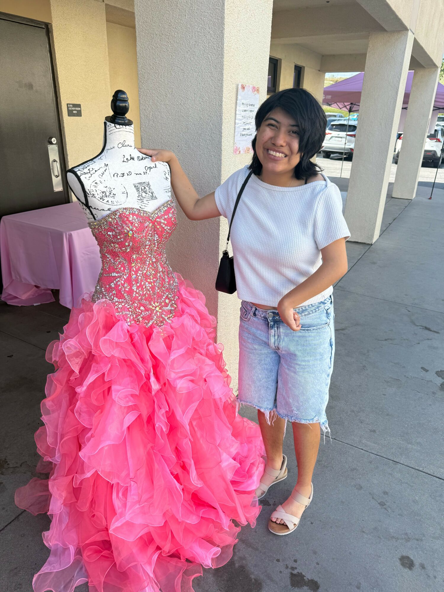Young girl in casual clothes stands next to a pink, ruffled dress on a mannequin, indoors. She has a black bag.
