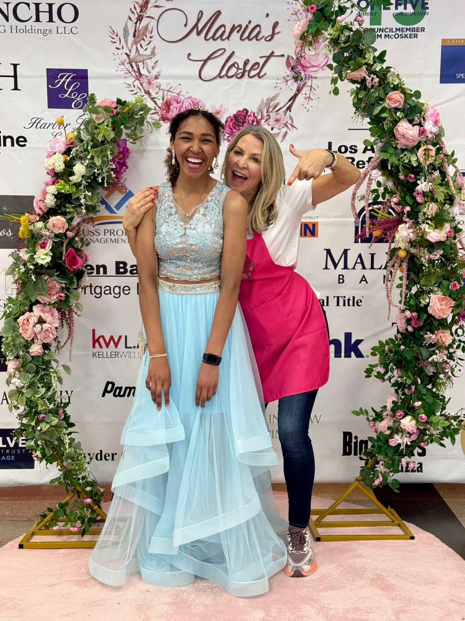 Two women smiling and posing inside a floral arch at an event, one in a light blue dress, the other in a pink apron and jeans.
