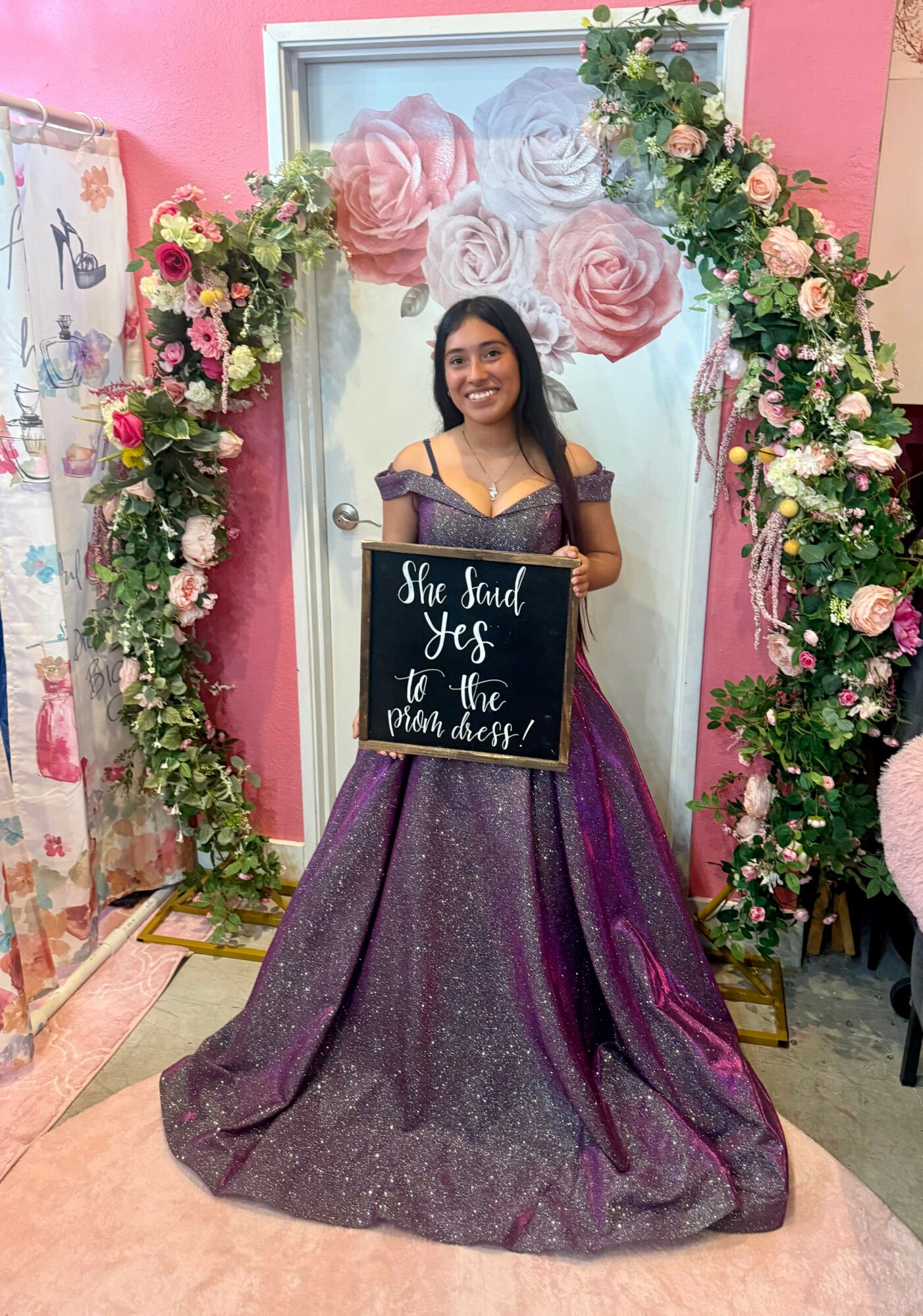 Young woman in purple gown holding a sign, standing in front of a floral arch and pink wall, smiling.