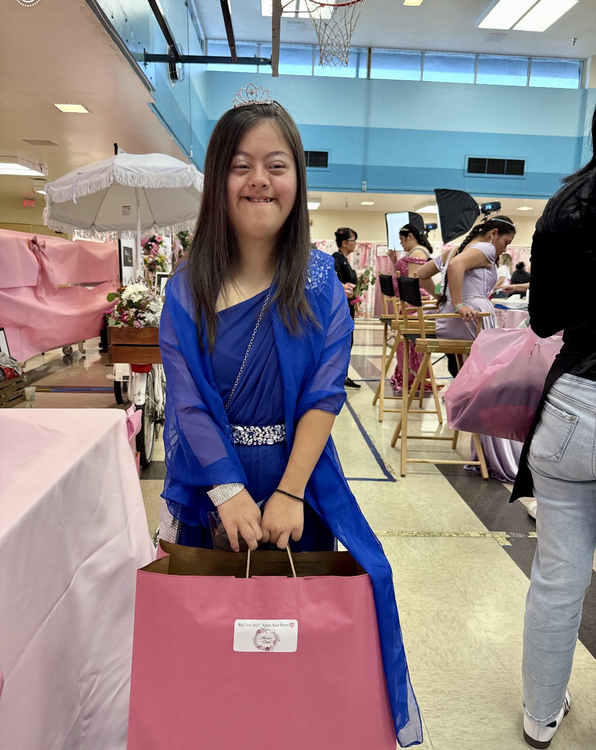 Young woman in blue dress holding a pink gift bag, smiling indoors at an event with tables and people.