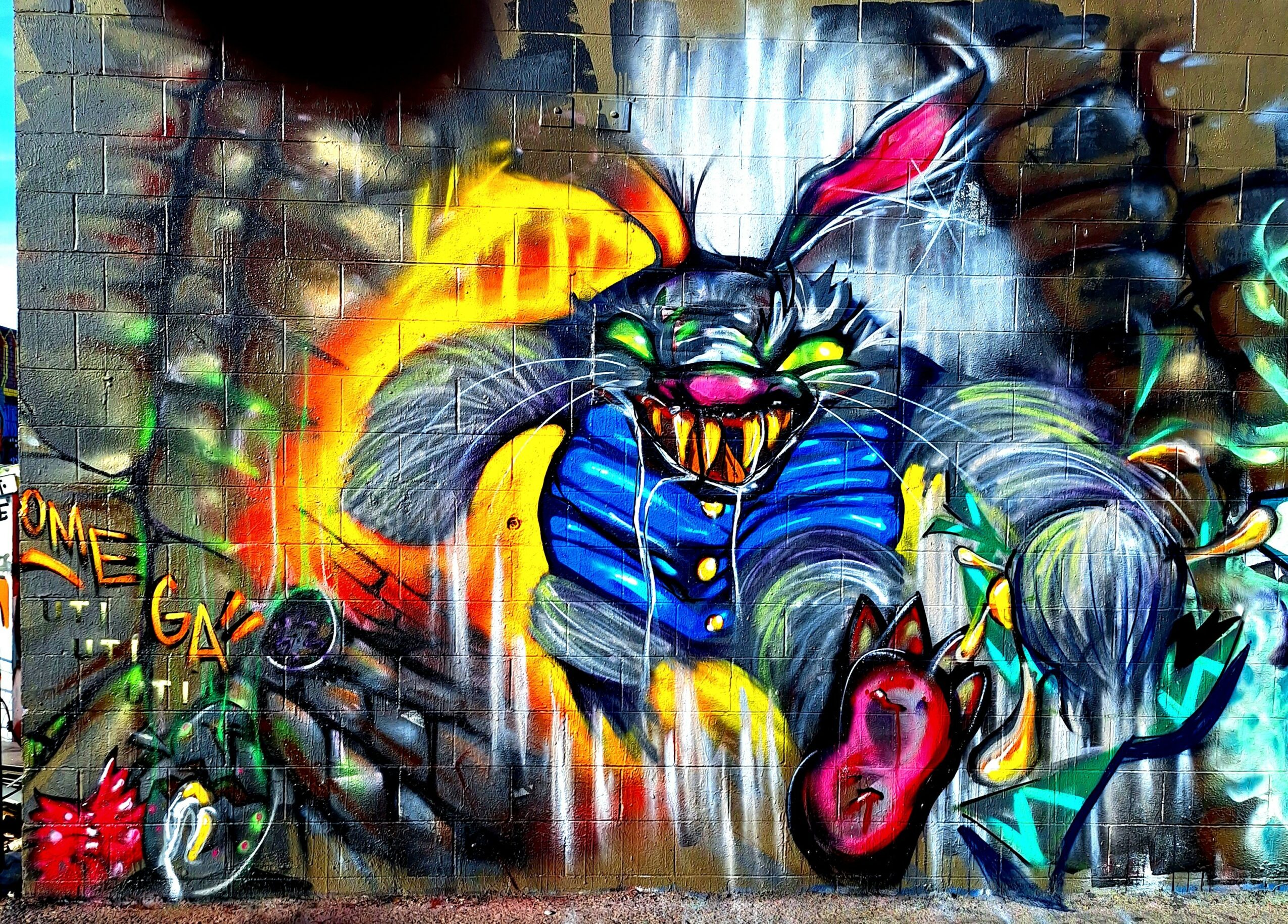 Colorful graffiti of a mischievous cat with sharp teeth, wearing a blue jacket, surrounded by spray paint cans and graffiti elements.