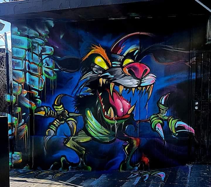 Colorful graffiti of a fierce, cartoonish cat with sharp teeth, glowing eyes, and exaggerated claws, on a dark background.
