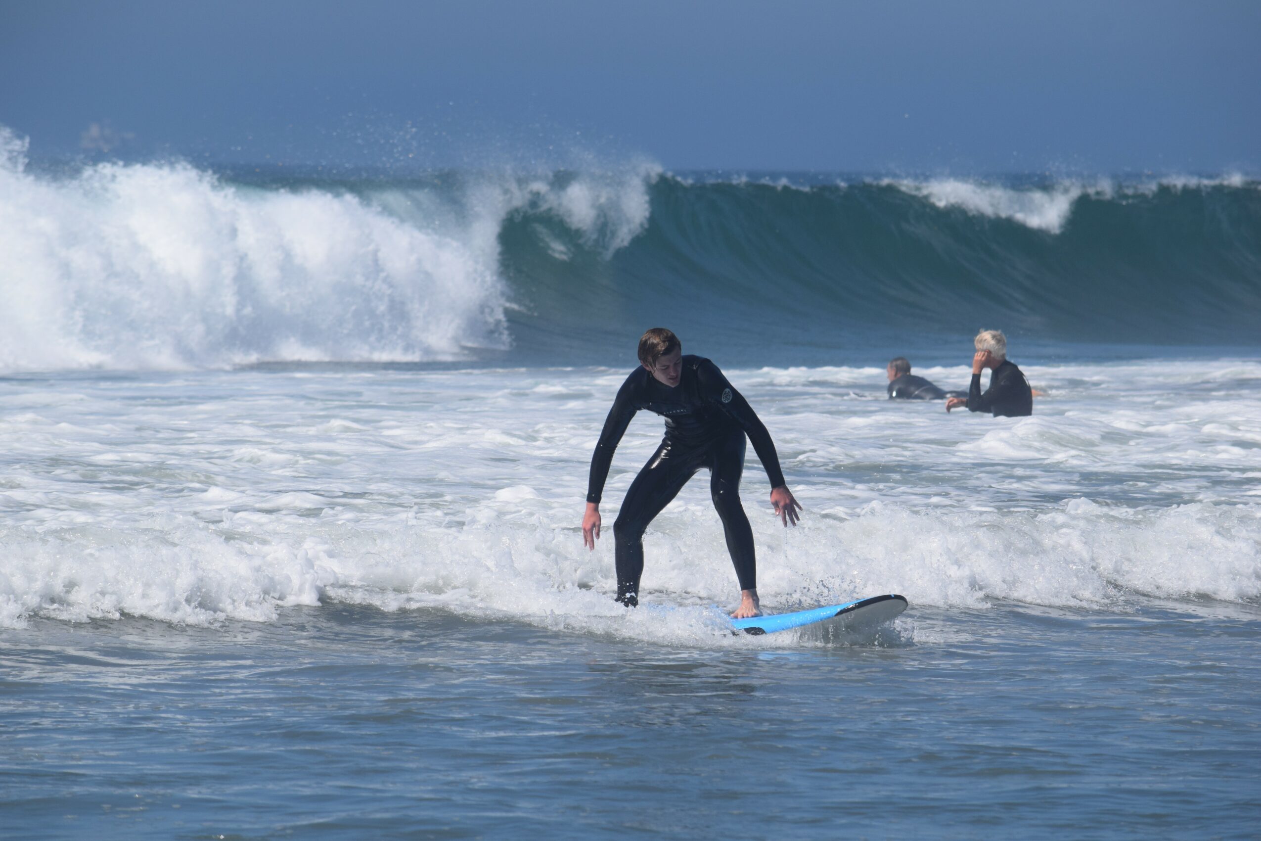 Person in wetsuit balancing on surfboard in ocean waves with two other surfers in background, large wave approaching.