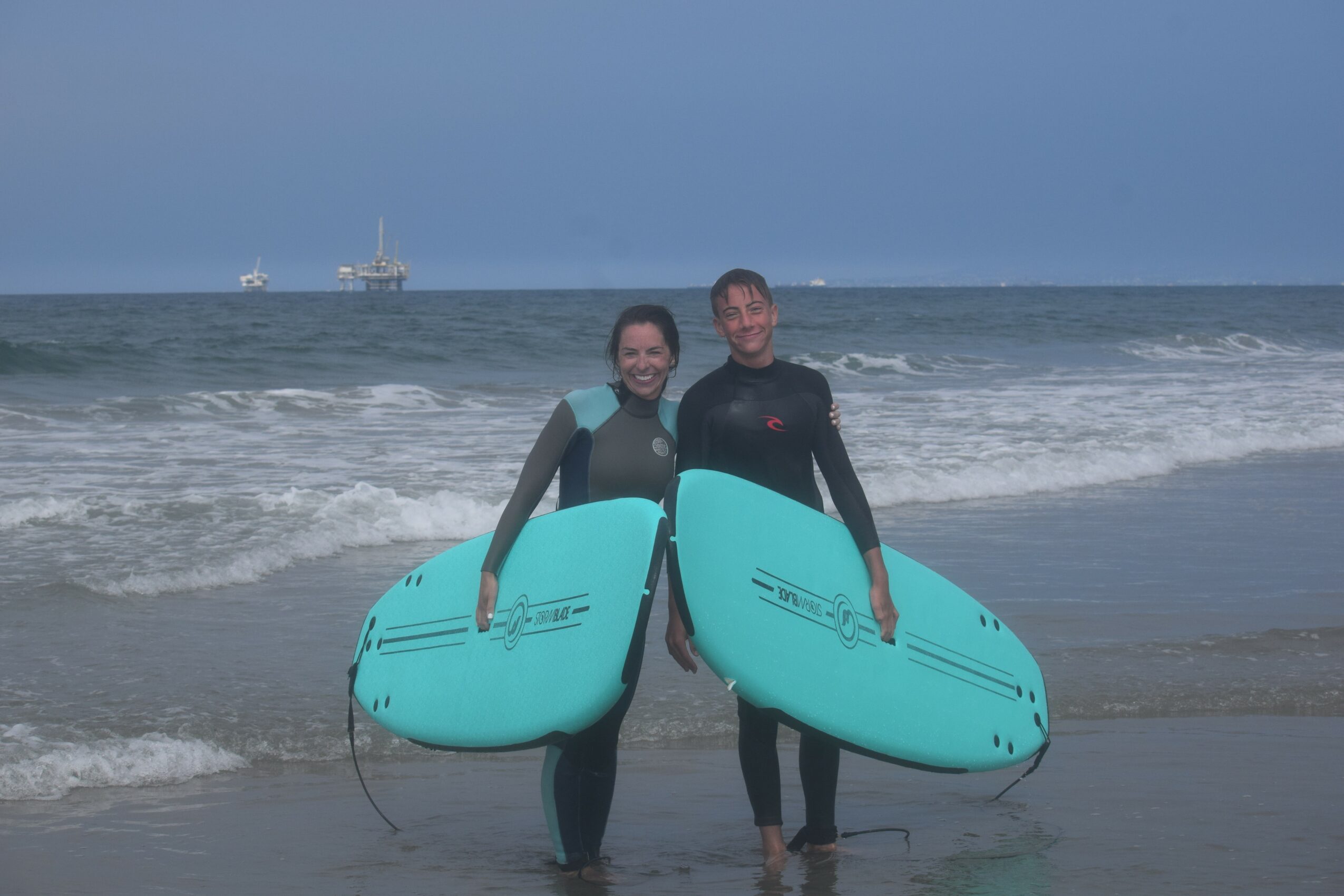 Two people in wetsuits holding surfboards on the beach with ocean waves behind them.
