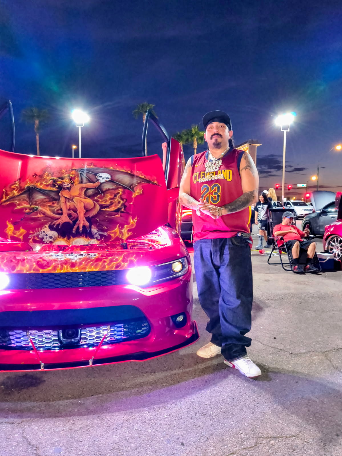 Person standing next to a pink car with a dragon design, in a parking lot during evening, illuminated by bright lights.