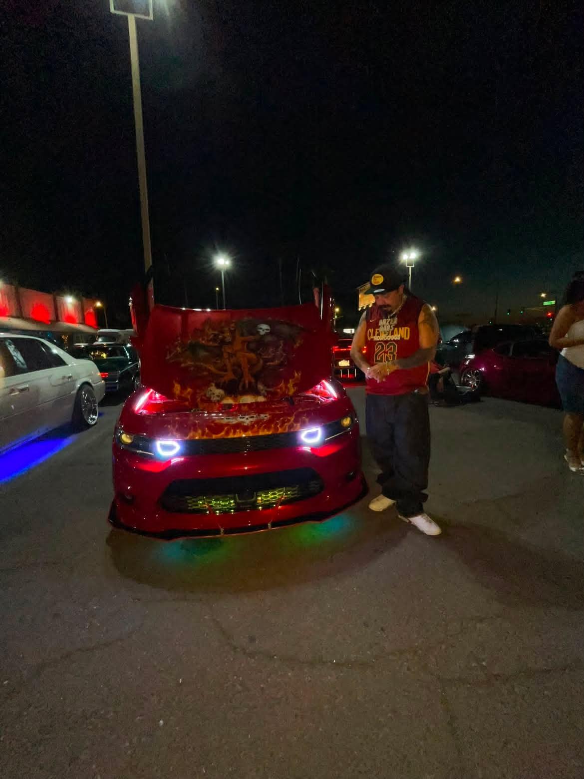 Person in a vest and cap standing next to a red car with an open hood in a parking lot at night, illuminated by streetlights.