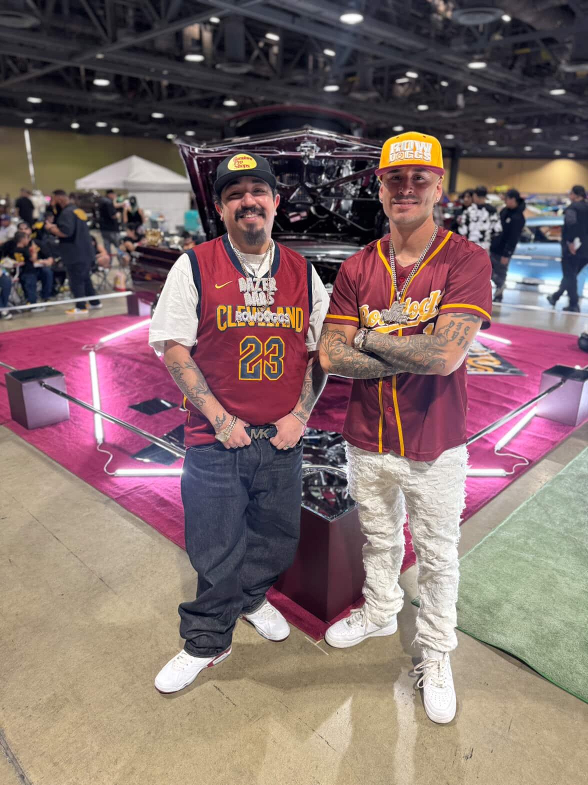 Two men stand in front of a car at an indoor event, smiling and wearing casual clothing and hats.