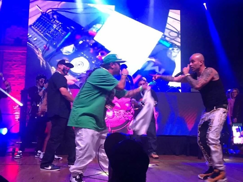 Two men are performing on stage with a DJ in the background, one holding a microphone and the other reaching out.