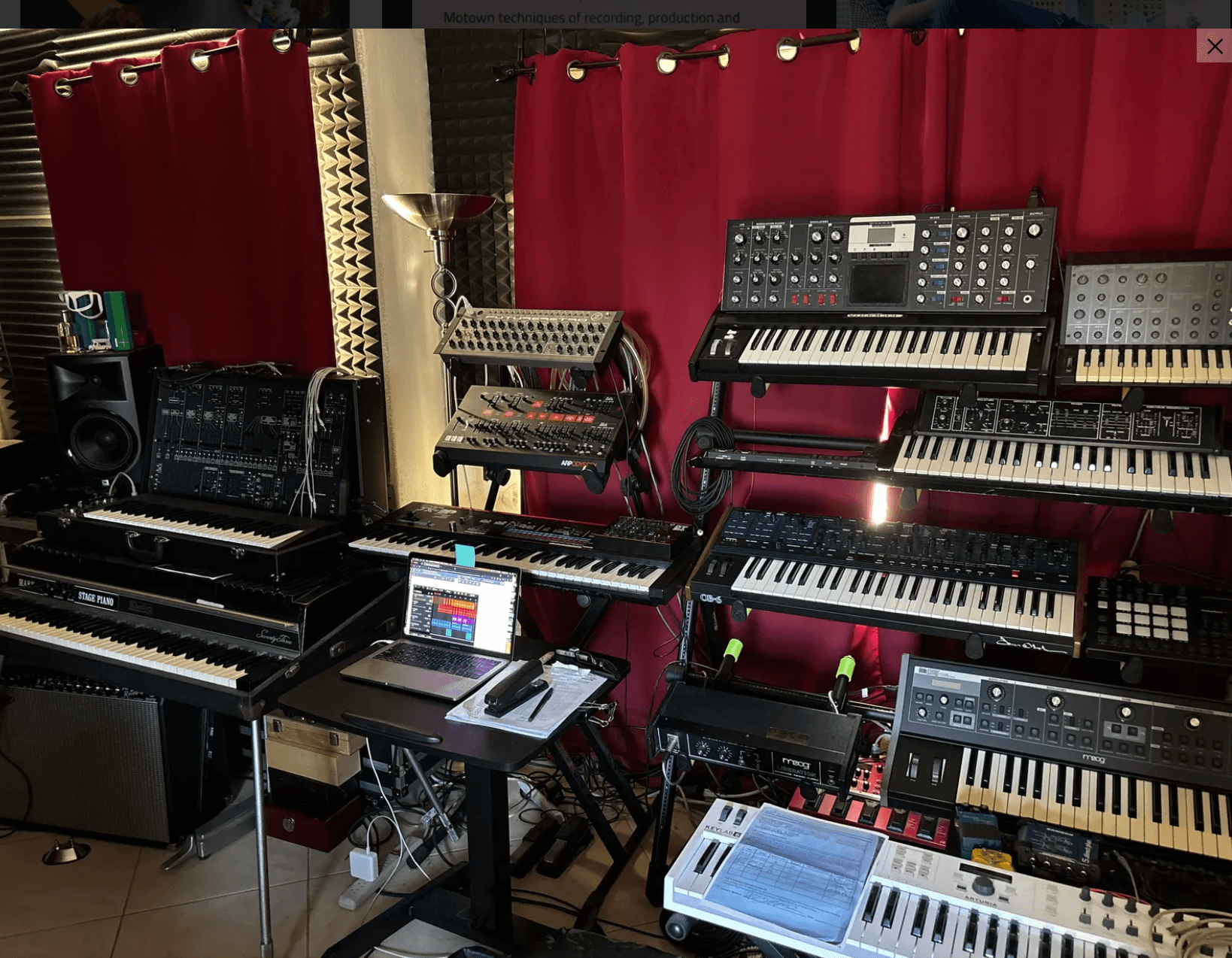Music studio with multiple keyboards, synthesizers, and electronic equipment arranged in front of red curtains.
