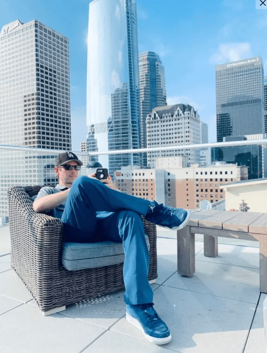 Person sitting on a wicker chair on a rooftop with city skyscrapers in the background, wearing sunglasses and casual clothing.