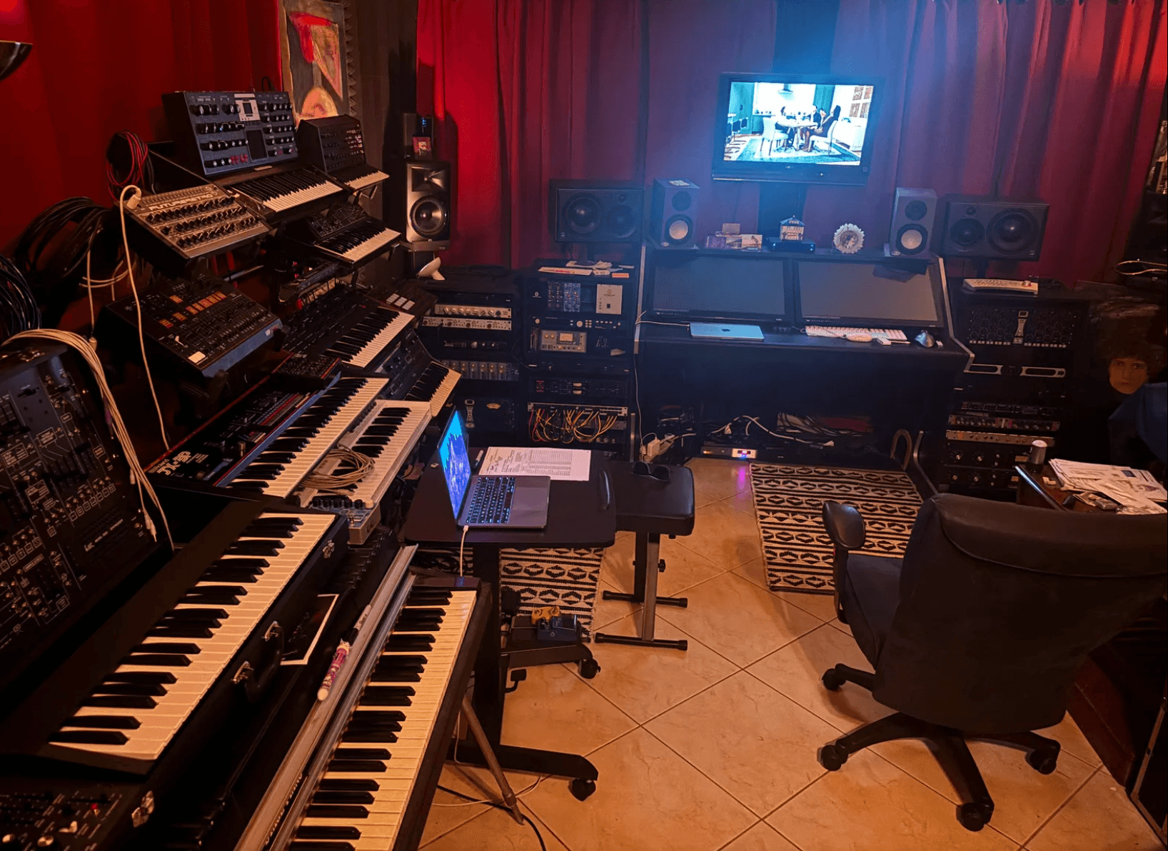 Music studio with keyboards, mixing equipment, speakers, and a computer monitor on a desk, red curtains in the background.
