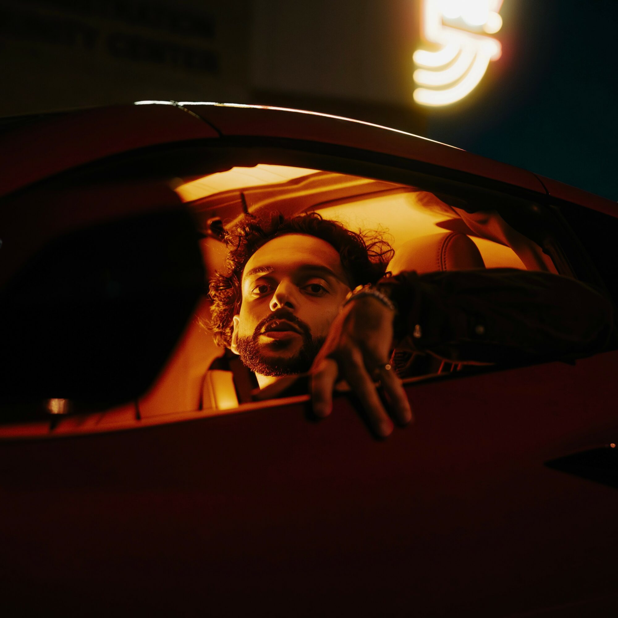 Man with curly hair and beard looking out of car window at night, illuminated by warm interior light.