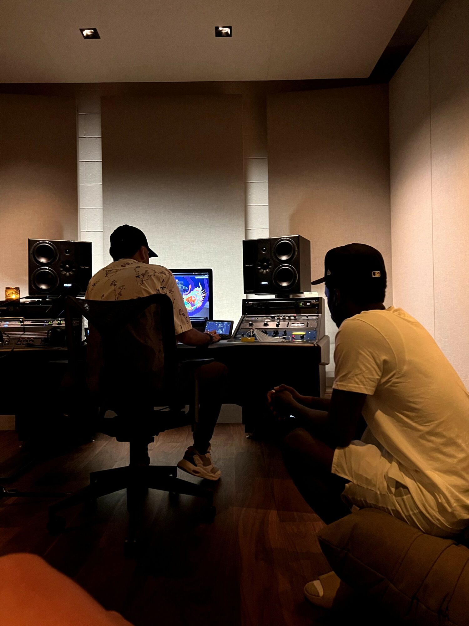 Two people in a recording studio, one working at a computer and audio equipment, the other sitting on the floor, facing the computer.