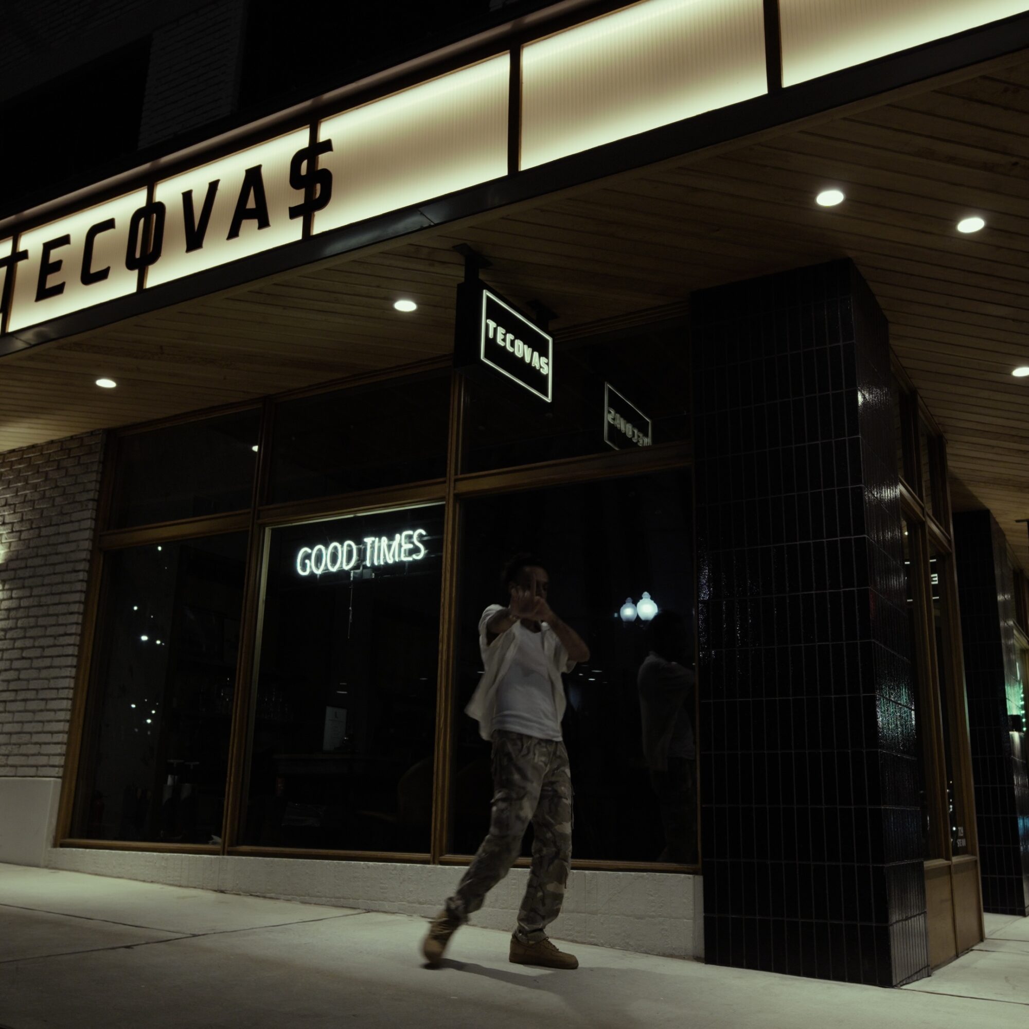 Person taking a photo outside a store with illuminated signs, large glass windows, and a black tiled pillar.