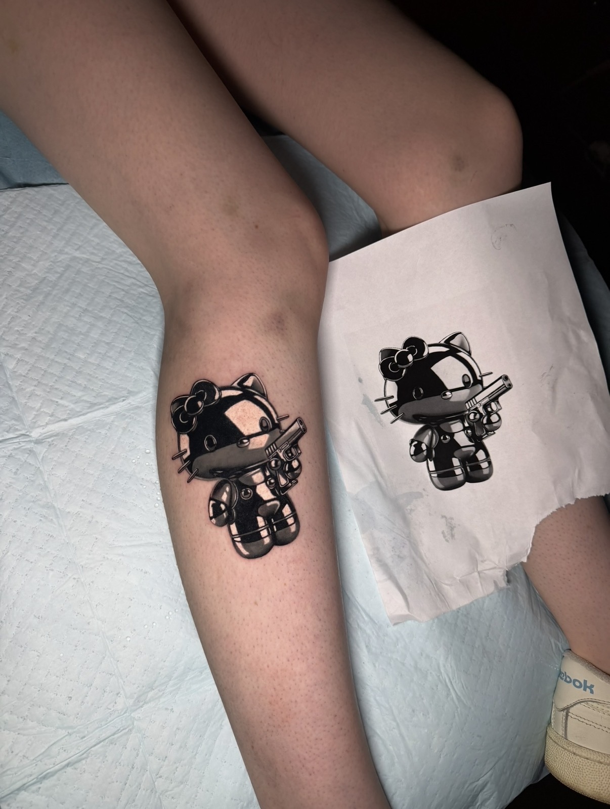 Child's leg with a tattoo of a cartoon character, next to a paper with the same tattoo design, on a bed.