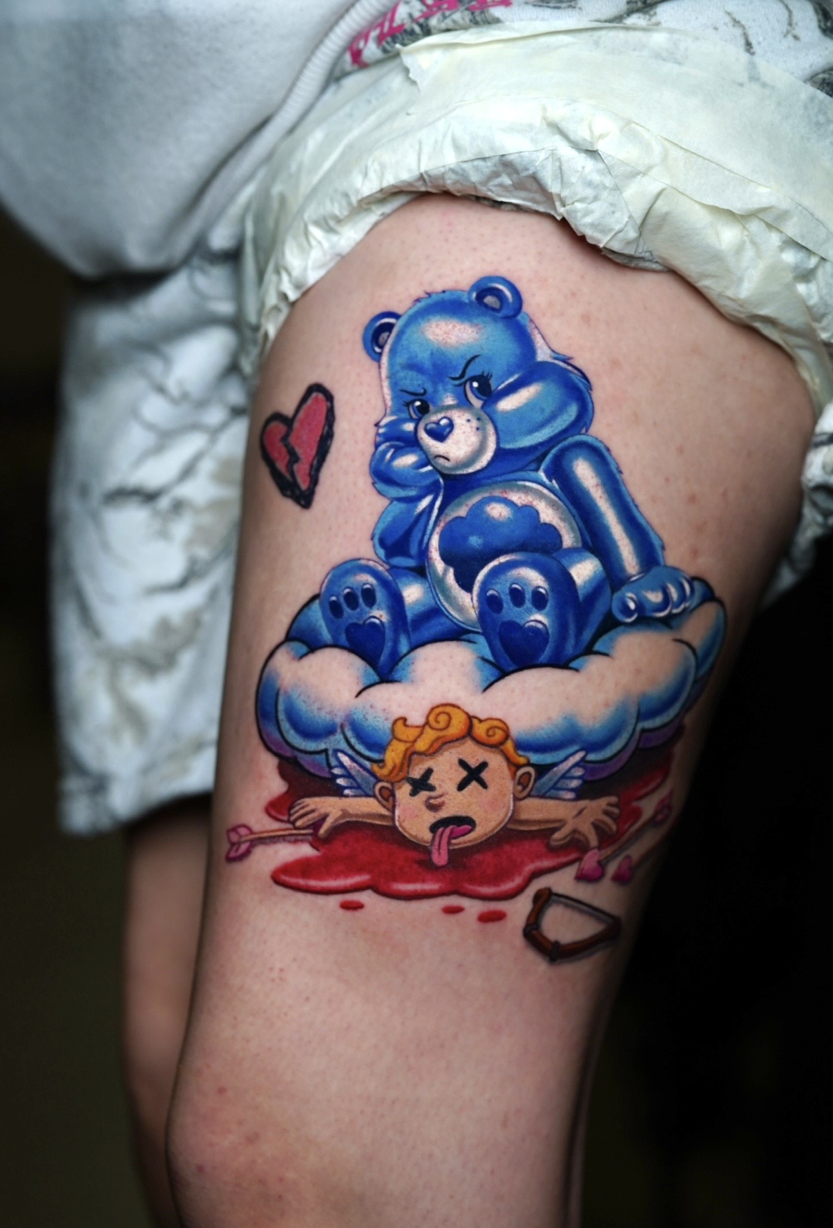 Tattoo of a blue teddy bear sitting on a cloud with a small, dead, cartoonish animal below, and a red heart nearby.