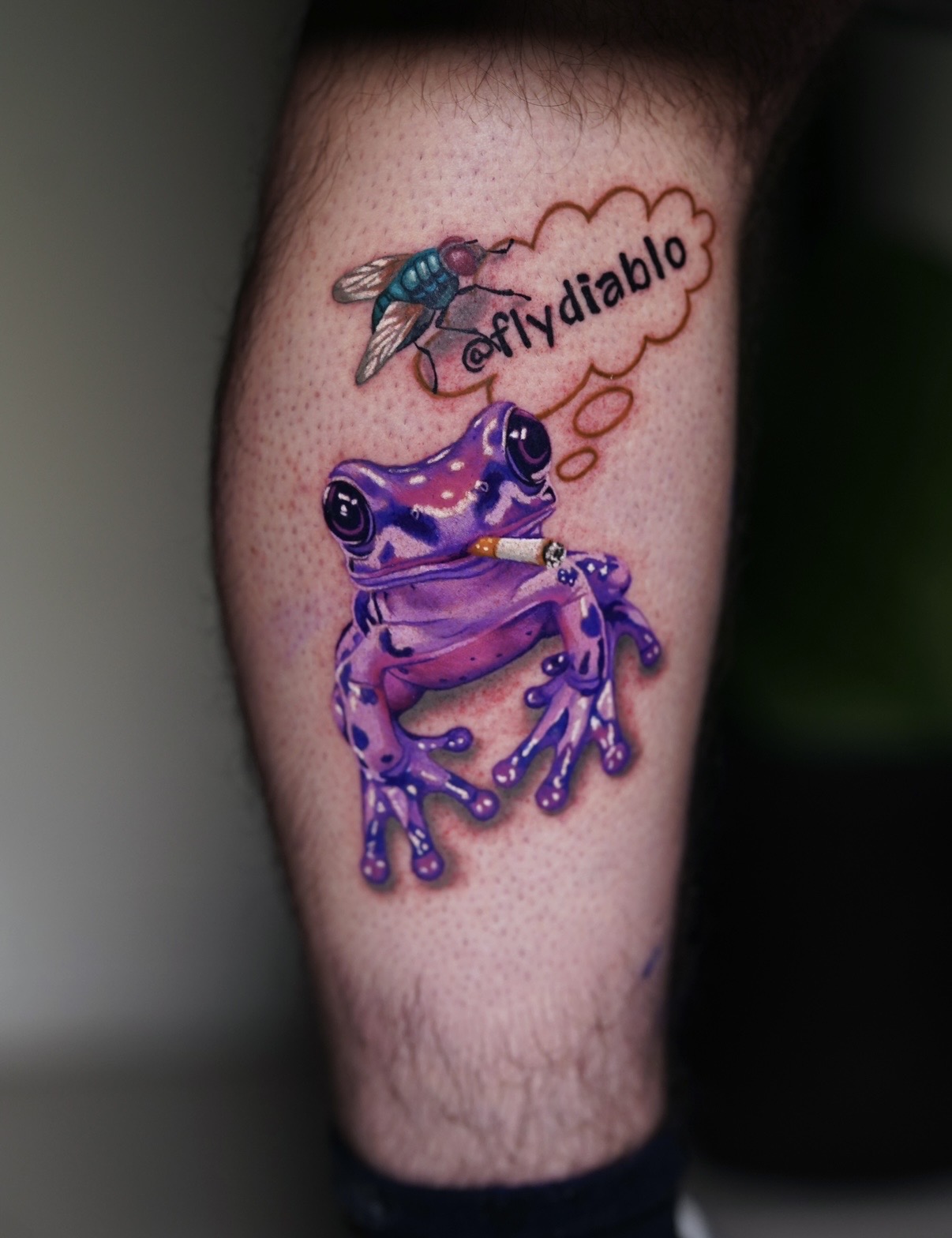 Tattoo of a purple frog with a speech bubble containing '@fydialbo' and a flying insect above it.