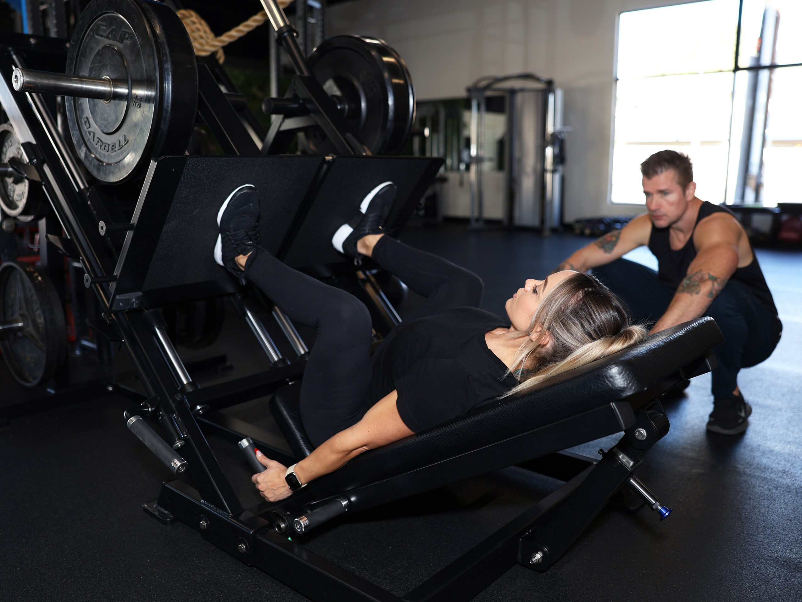 A woman lies on a leg press machine in a gym, with a trainer nearby observing her exercise.