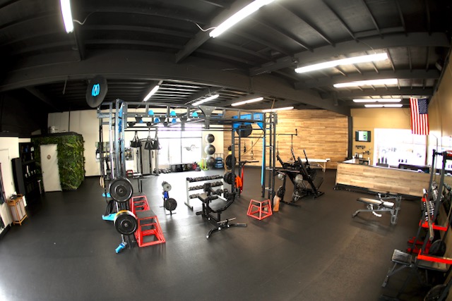 Gym with weightlifting equipment, benches, and a wooden wall, illuminated by ceiling lights, with an American flag on the wall.