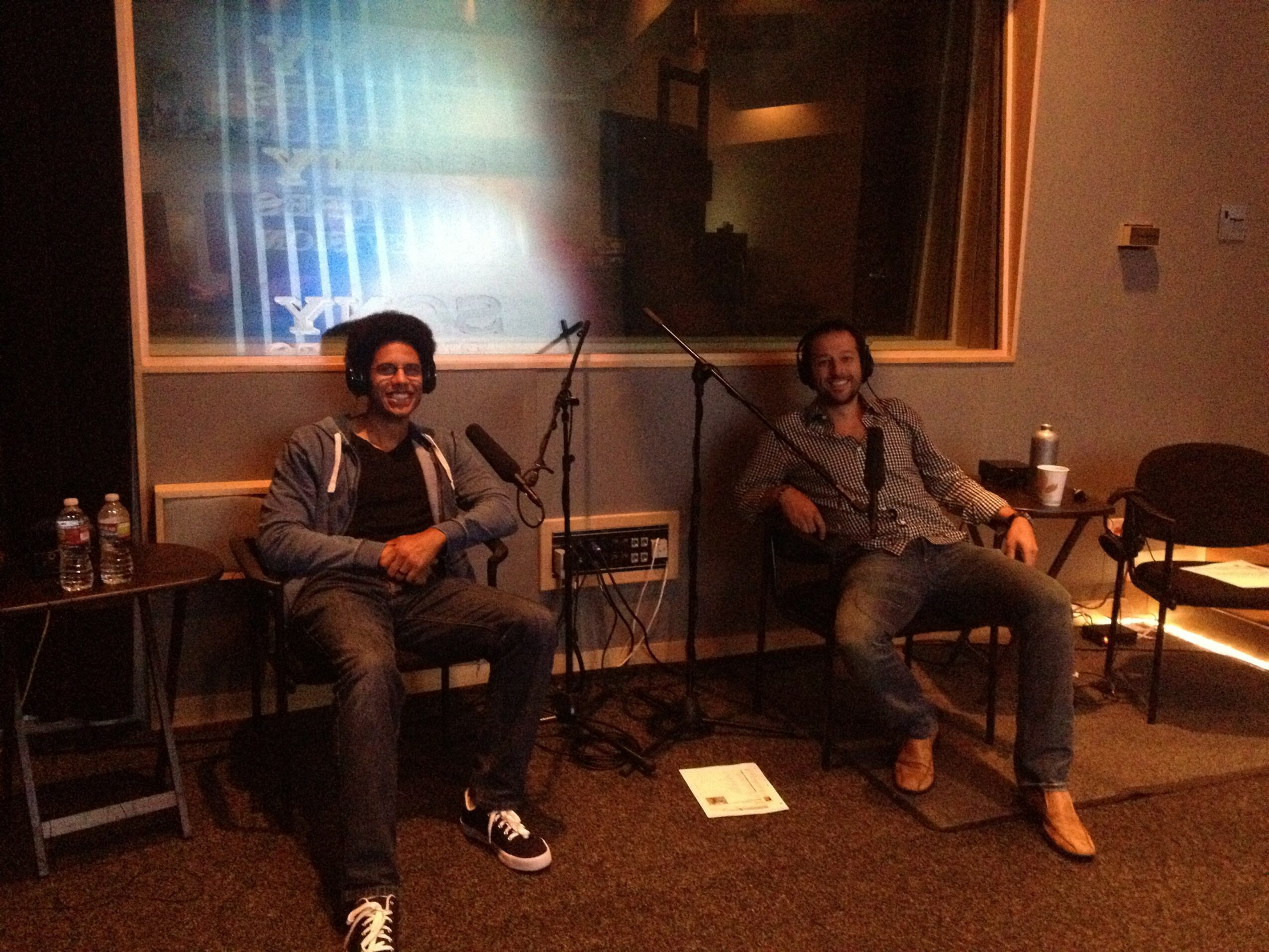 Two people sit in a recording studio with microphones, one smiling and the other relaxed, in a dimly lit room.
