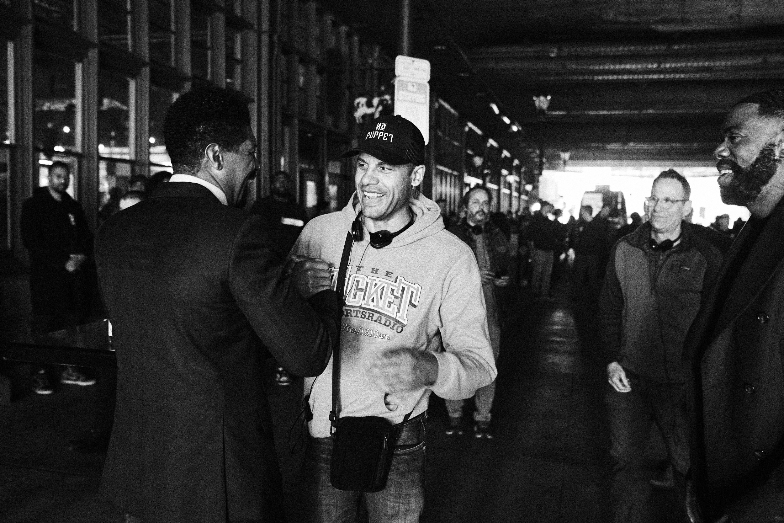 Three men are engaged in conversation in an indoor setting with a group of people in the background, some wearing glasses and jackets.
