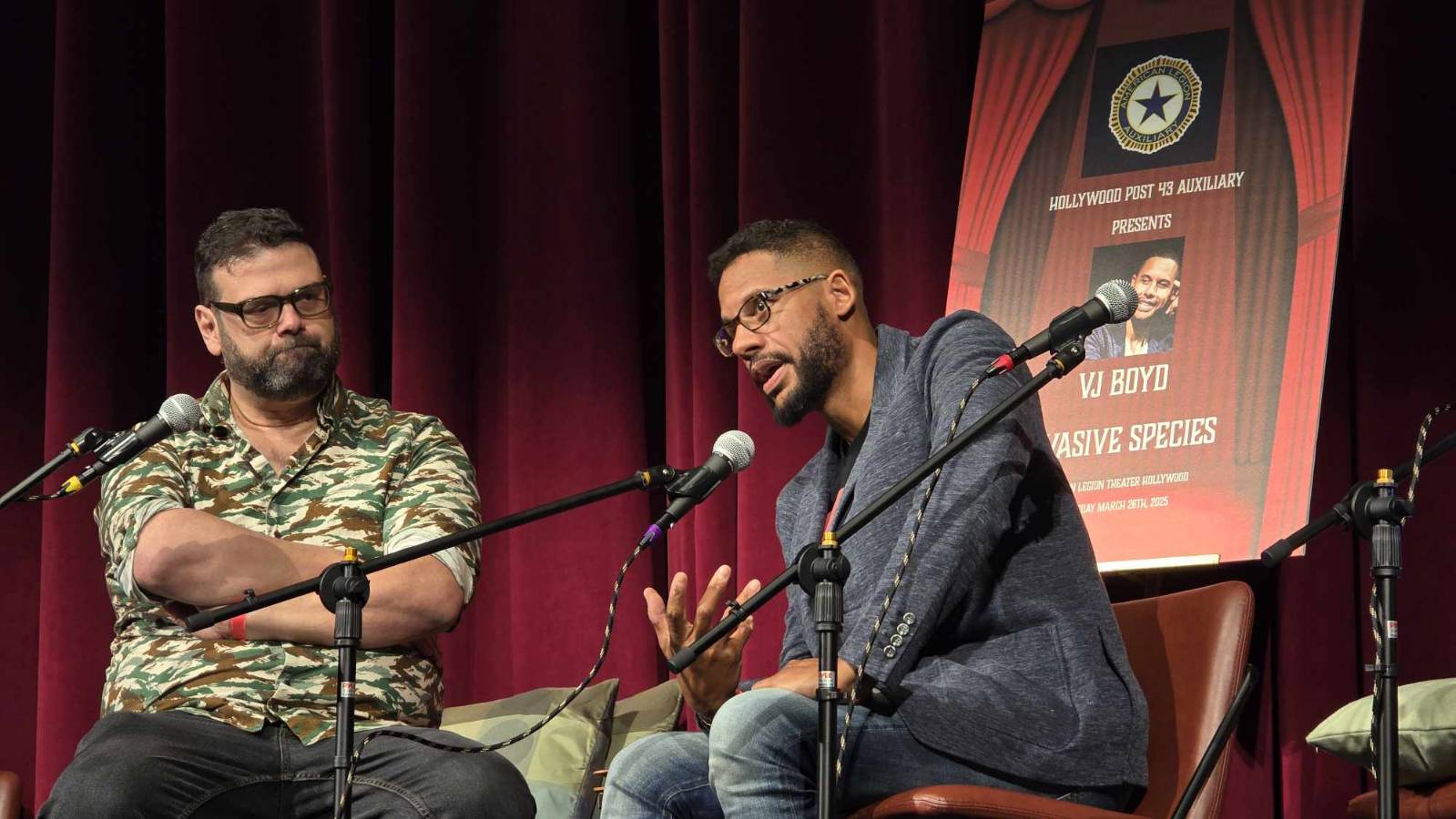 Two men sit on stage with microphones, engaged in conversation, in front of a red curtain and a poster, one gesturing.