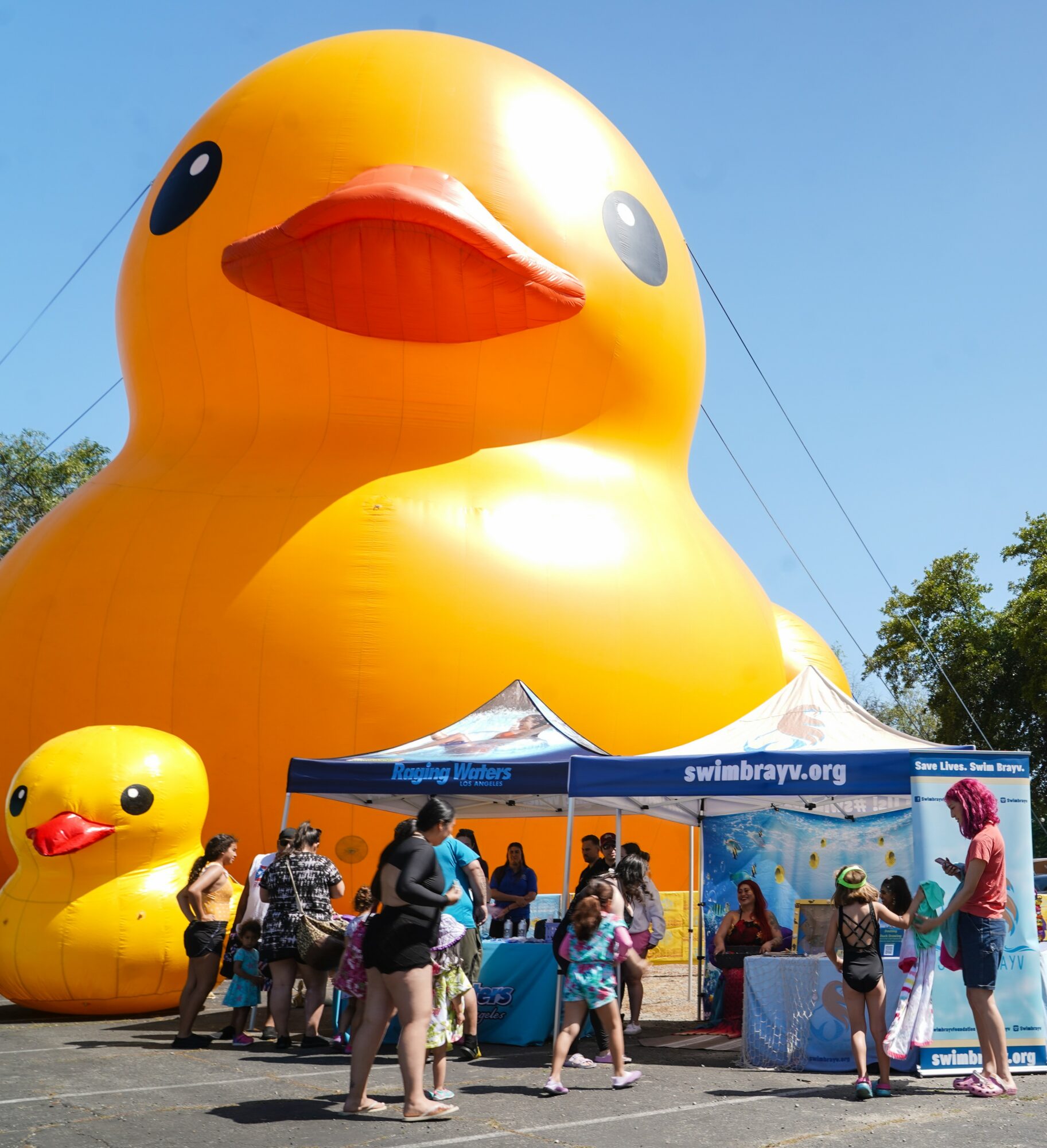 Large inflatable yellow rubber duck with smaller duck and people gathered around a tent, outdoors on a sunny day.