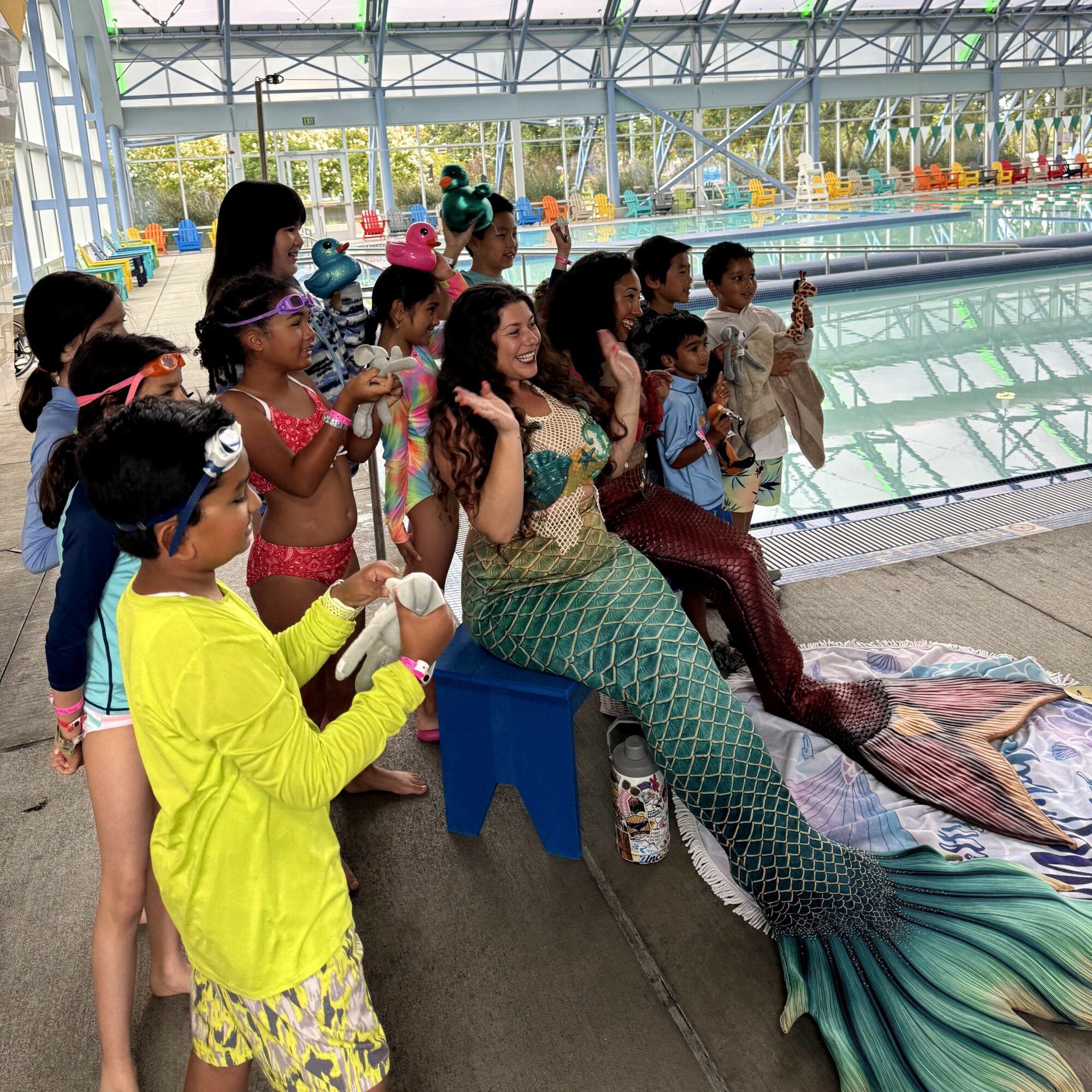 Group of children and a woman dressed as a mermaid sitting by an indoor pool, some children holding objects, bright environment.