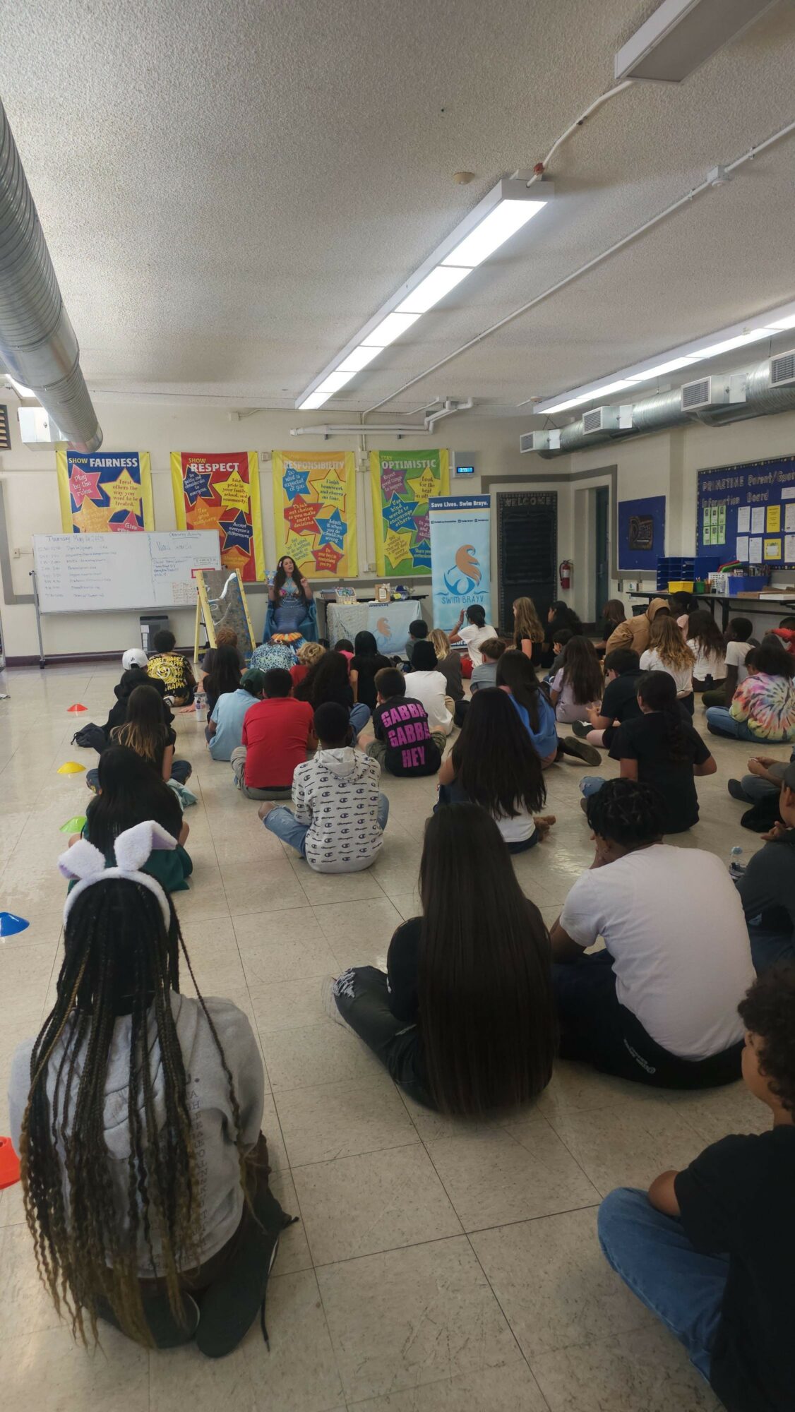 People standing and sitting in a crowded indoor space, possibly a waiting area or hallway, with colorful posters on the wall.