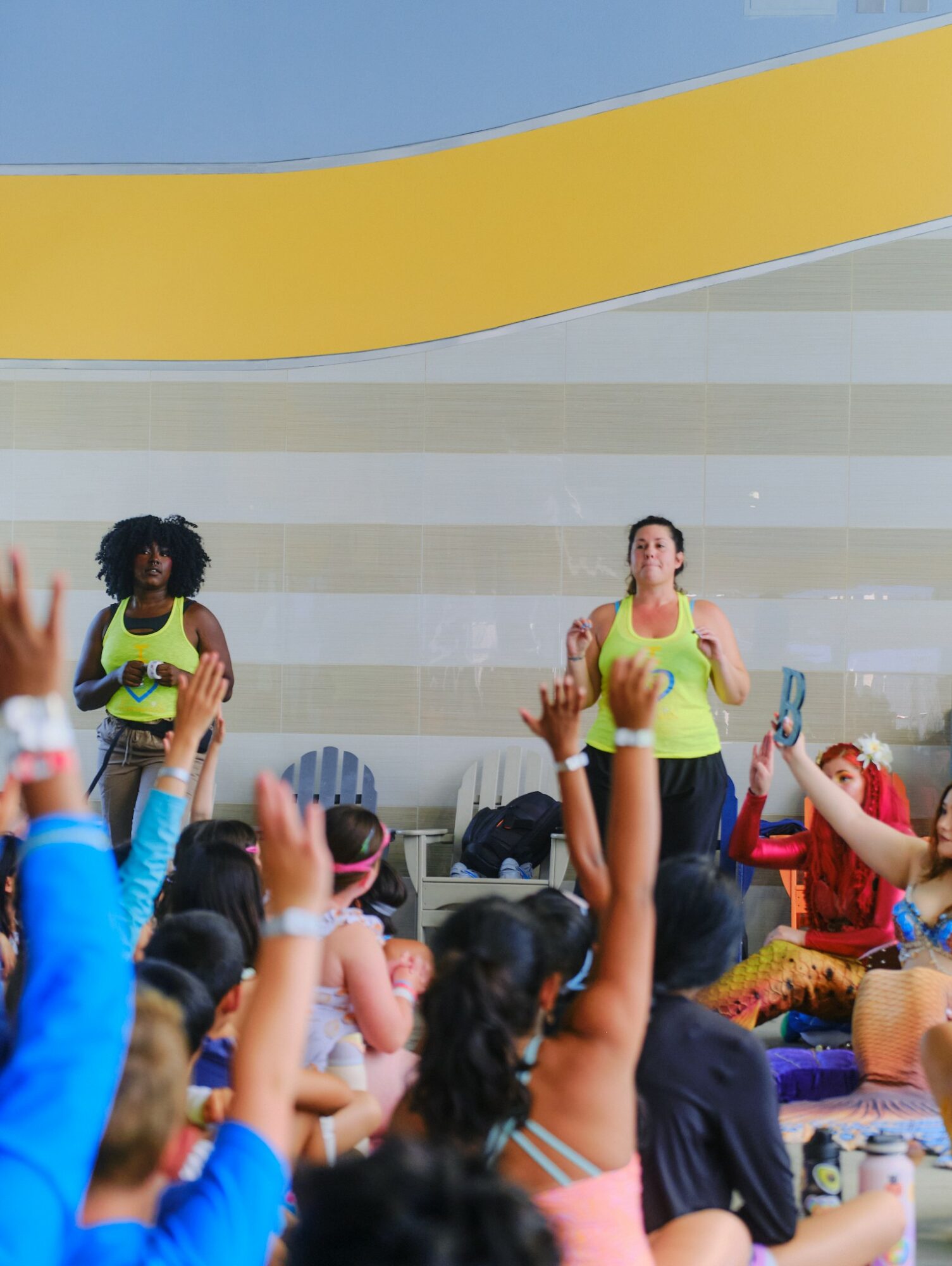 Two women in yellow tops lead a fitness class with children raising hands, in a colorful indoor setting.