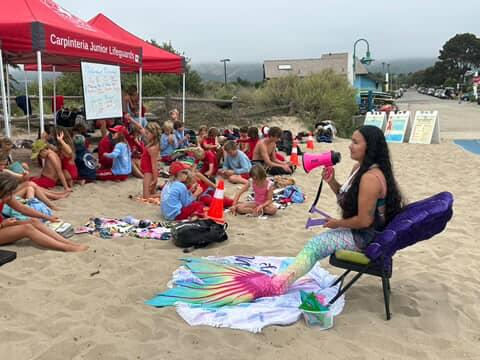 Group of children and adults sitting on sand near a red canopy, with a woman in mermaid costume sitting on a chair, holding a pink megaphone.