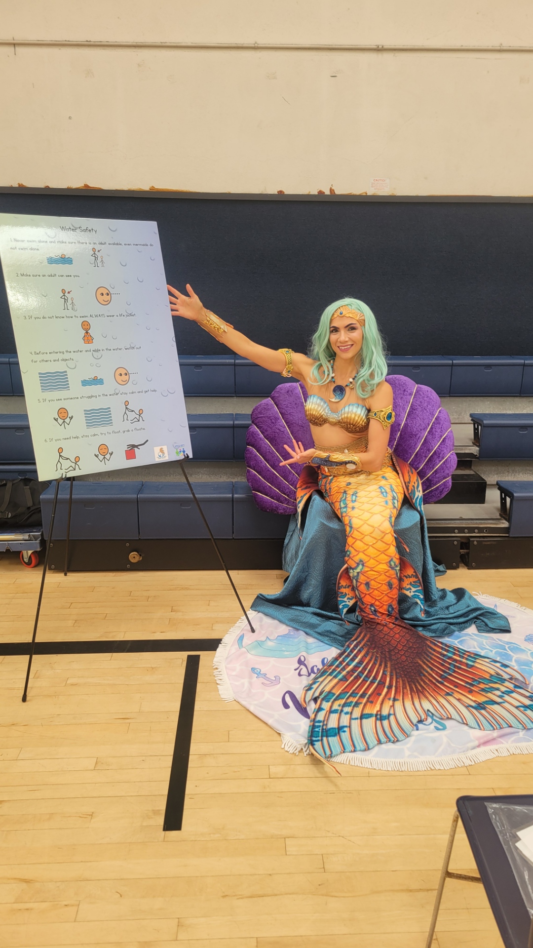 Person dressed as a mermaid with a purple shell-shaped chair, pointing at a display board in a gymnasium.
