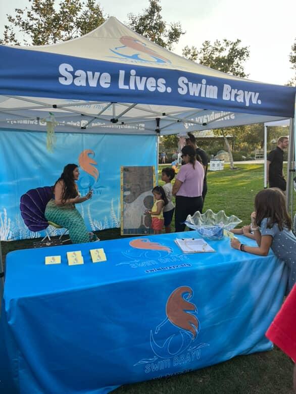 People gather under a blue tent with a banner that reads 'Save Lives. Swim Bravely.' at an outdoor event.