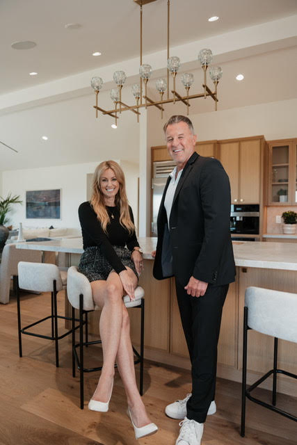 A woman and a man stand in a modern kitchen, smiling, with a chandelier overhead and bar stools nearby.