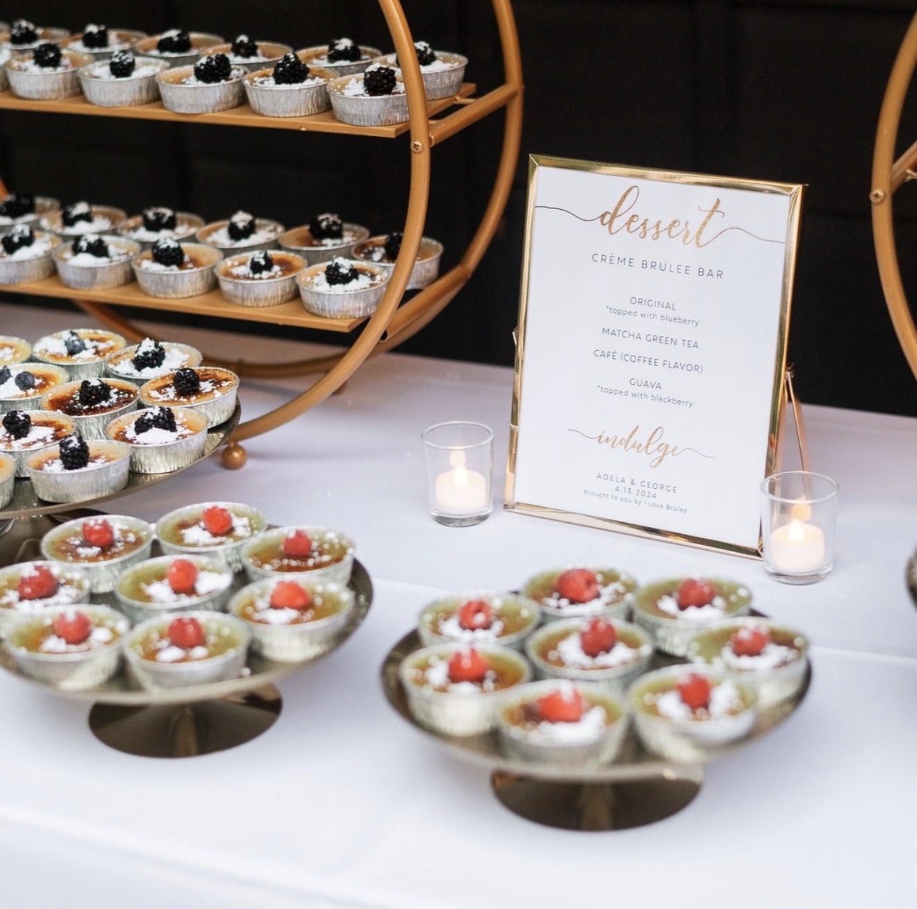 Dessert table with cupcakes and fruit-topped treats, a menu sign, and candles, set against a dark background.