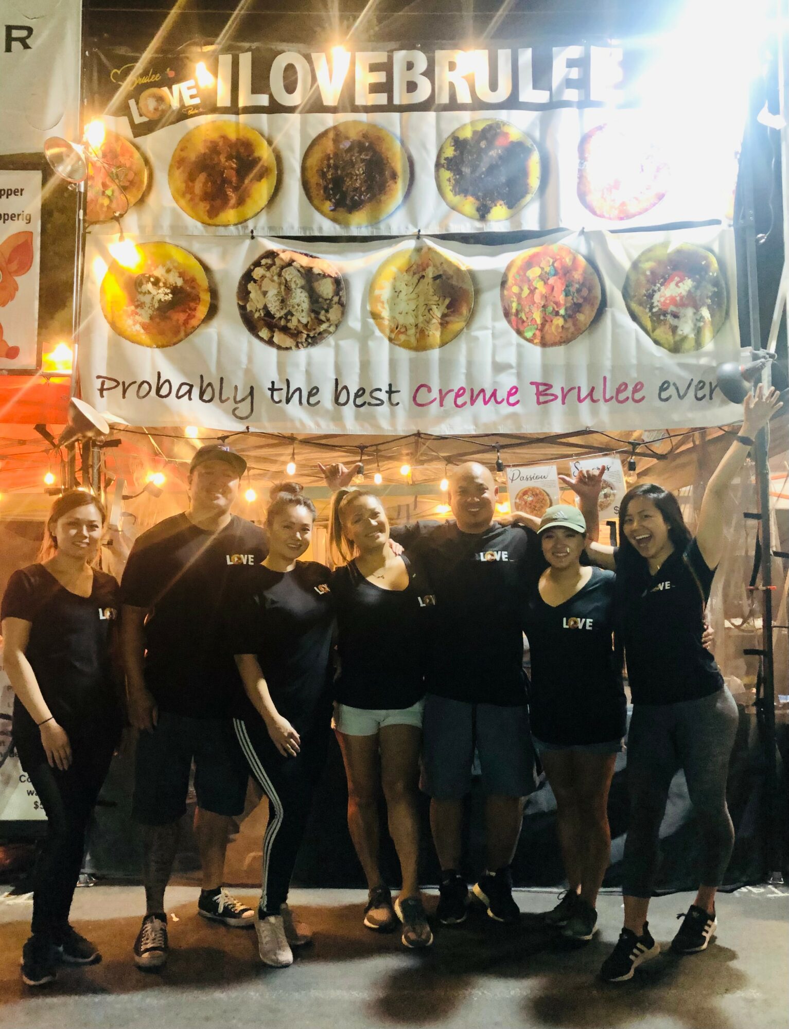 Group of people standing in front of a brightly lit food stall with a sign that reads 'LOVE BRULEE' and images of desserts, smiling and posing for the photo.