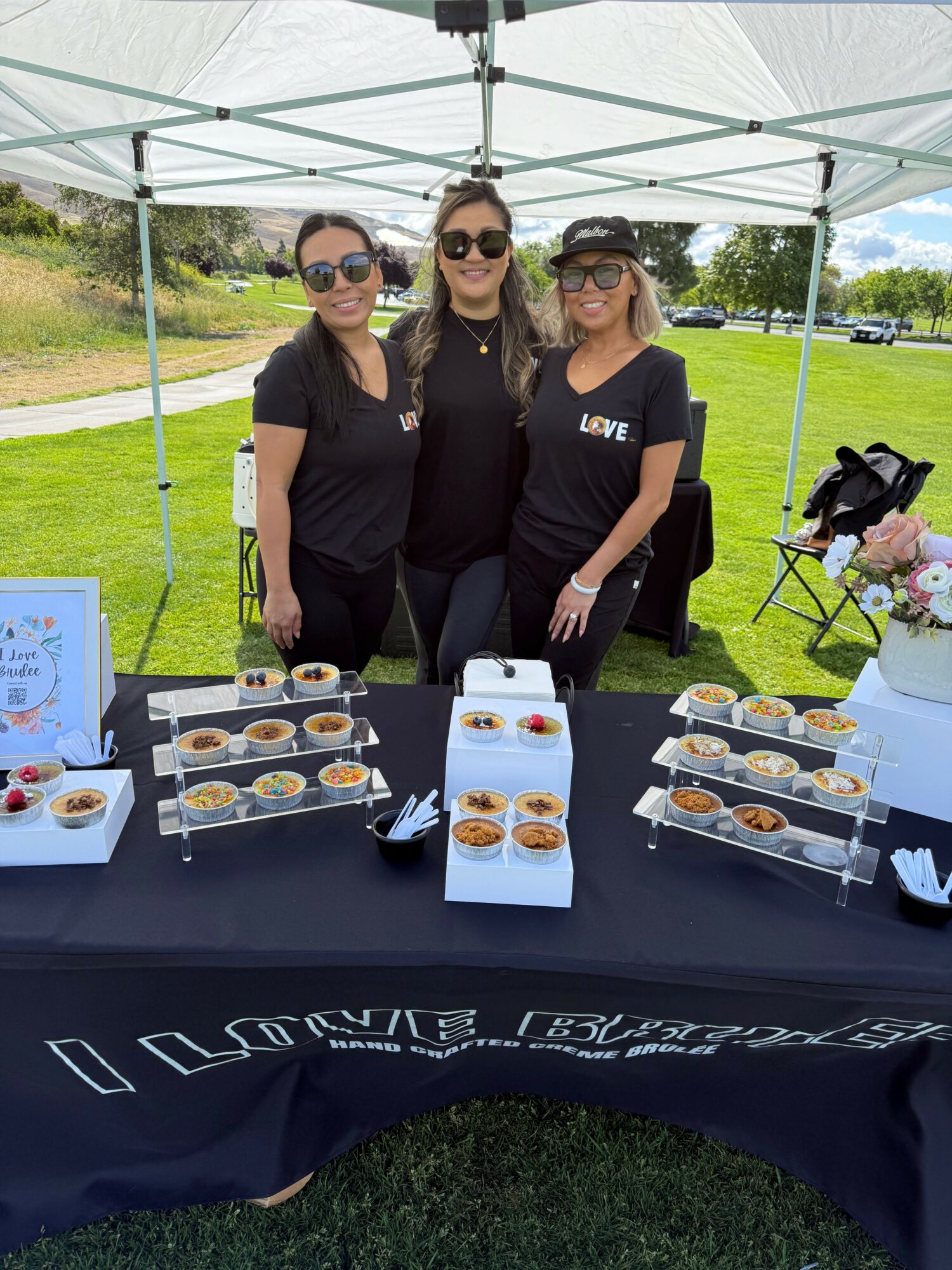 Three women in black clothing stand behind a table with food samples outdoors under a tent. Green grass and trees are in the background.