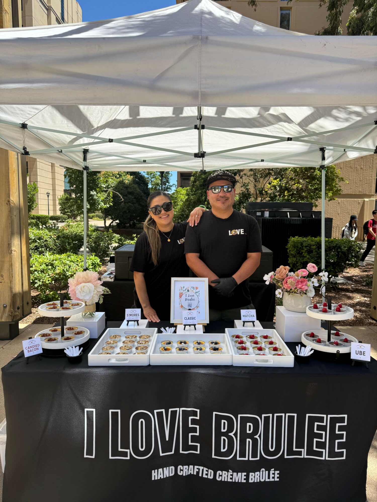 Two women stand behind a table with baked goods at an outdoor event, under a white canopy, with trees and buildings in background.