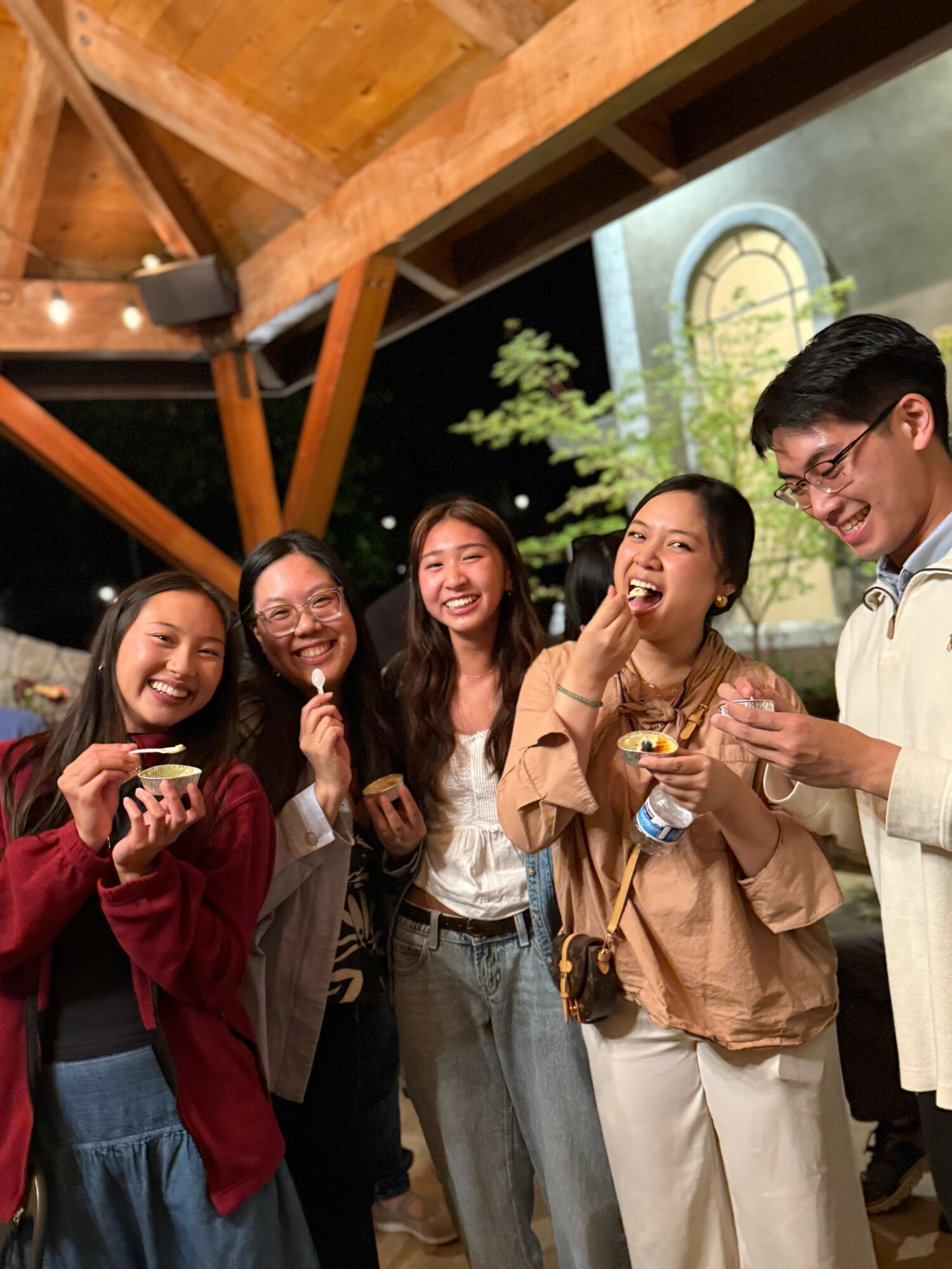 Group of five young women smiling and laughing together outdoors at night, under a wooden structure, with a building and tree in background.